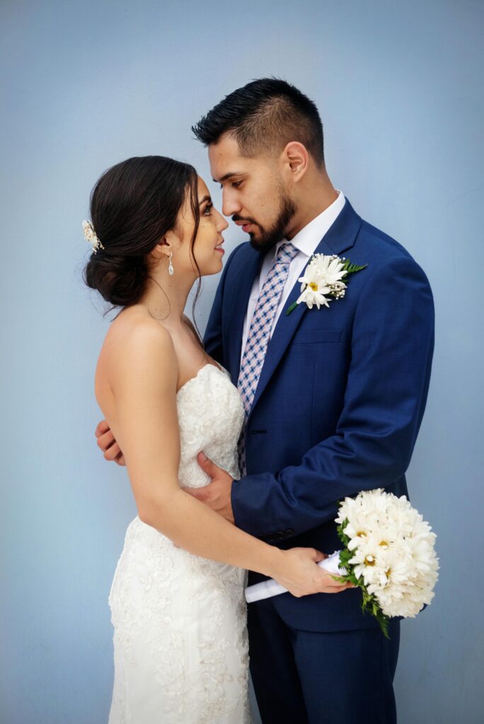 Bride and groom sharing a tender moment with bouquet against a blue background.