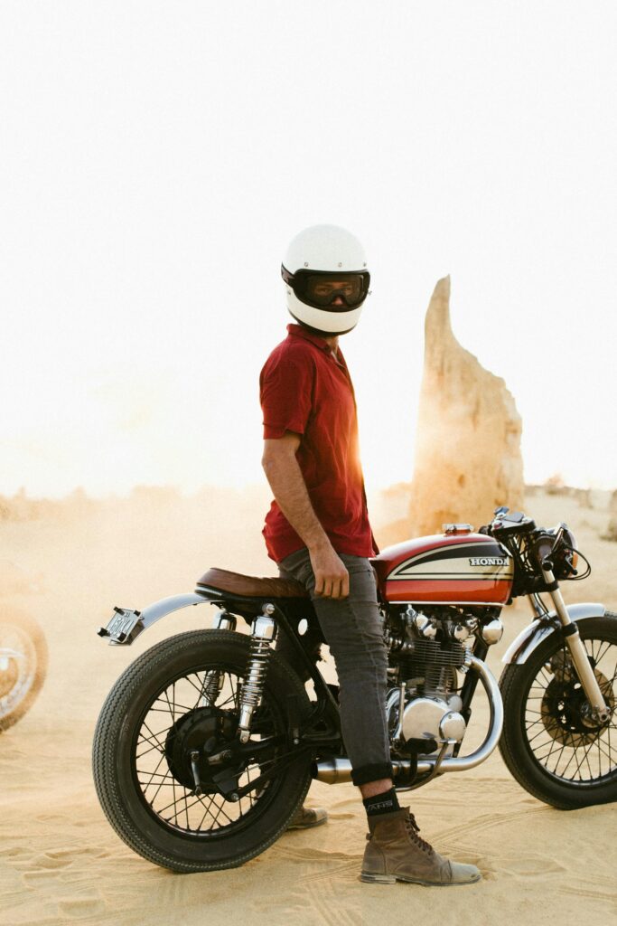 Full body of anonymous biker in casual clothing and helmet standing over motorcycle in desert under gray sky in sunlight